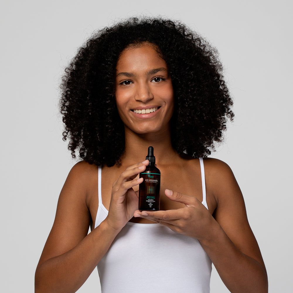 Eine Person mit lockigem Haar hält eine Flasche mit Pipette und lächelt in die Kamera.