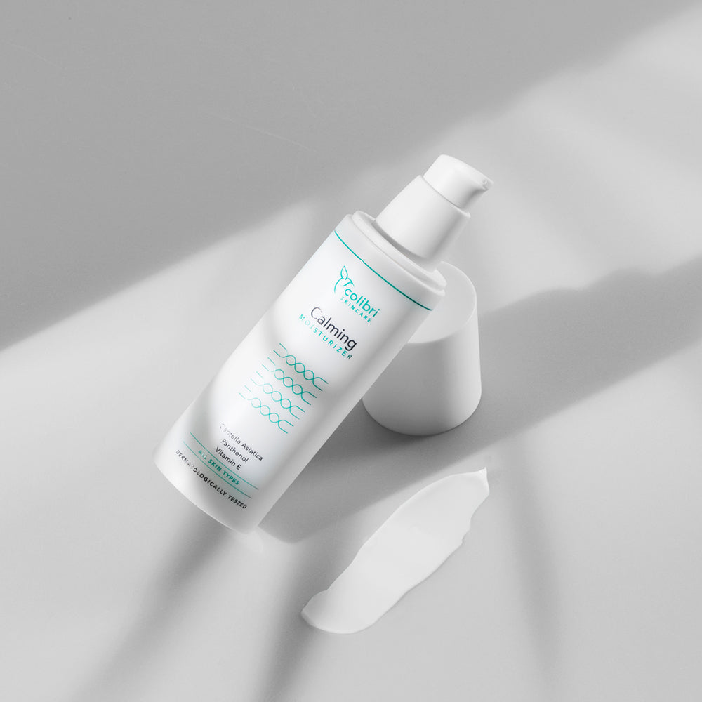 Weiße Flasche mit Feuchtigkeitscreme liegt auf grauem Hintergrund, daneben ist eine kleine Menge der Creme zu sehen.