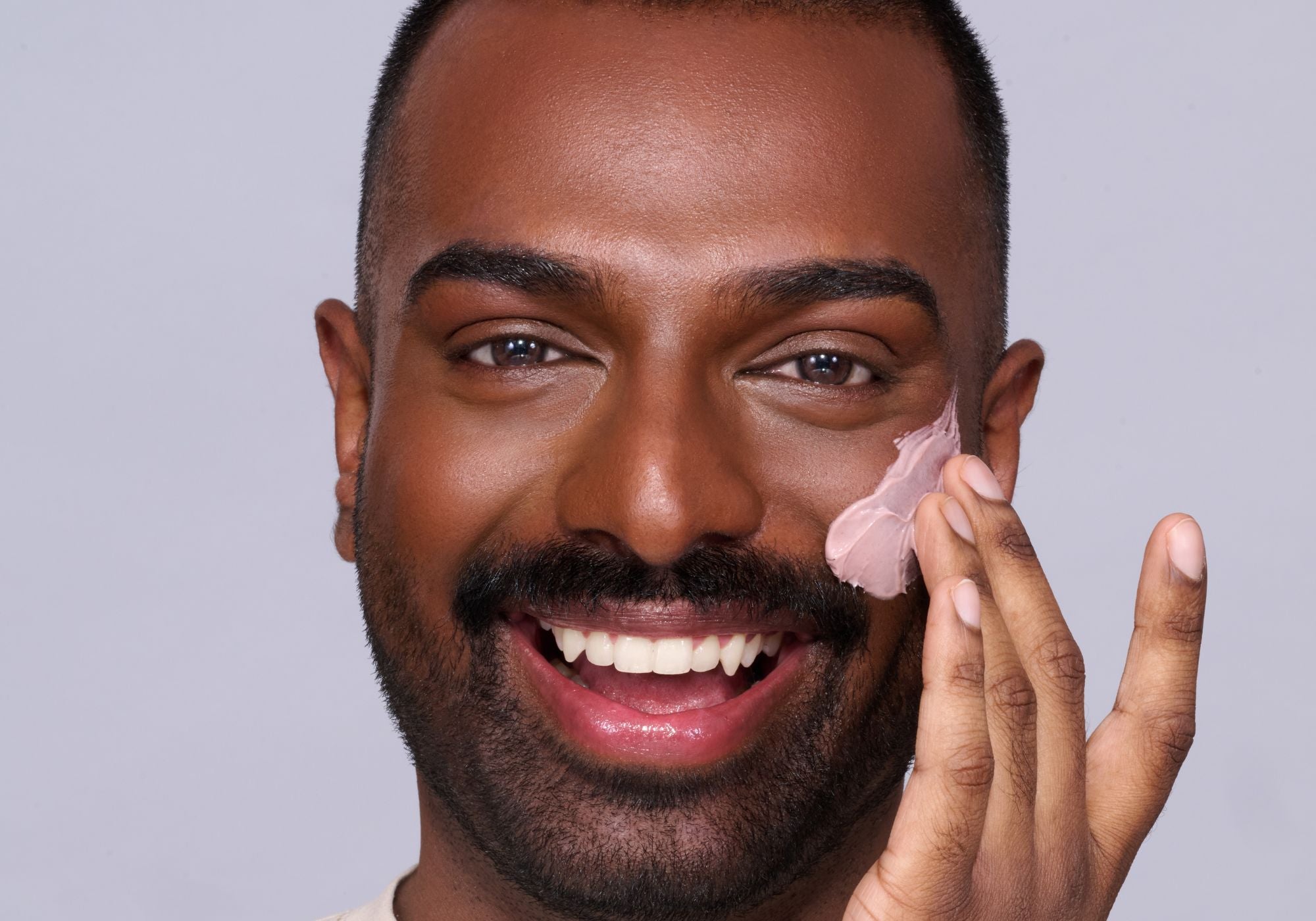 Lächelnder Mann trägt rosa Creme auf die Wange auf, vor grauem Hintergrund.