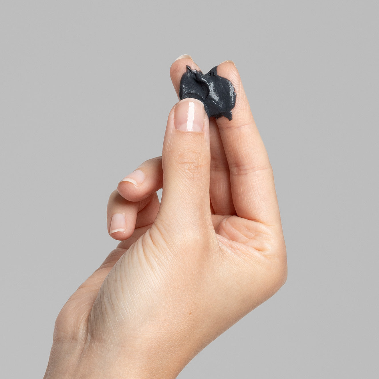 Hand hält einen kleinen Klumpen dunkelgrauer Knetmasse zwischen Daumen und Zeigefinger vor grauem Hintergrund.