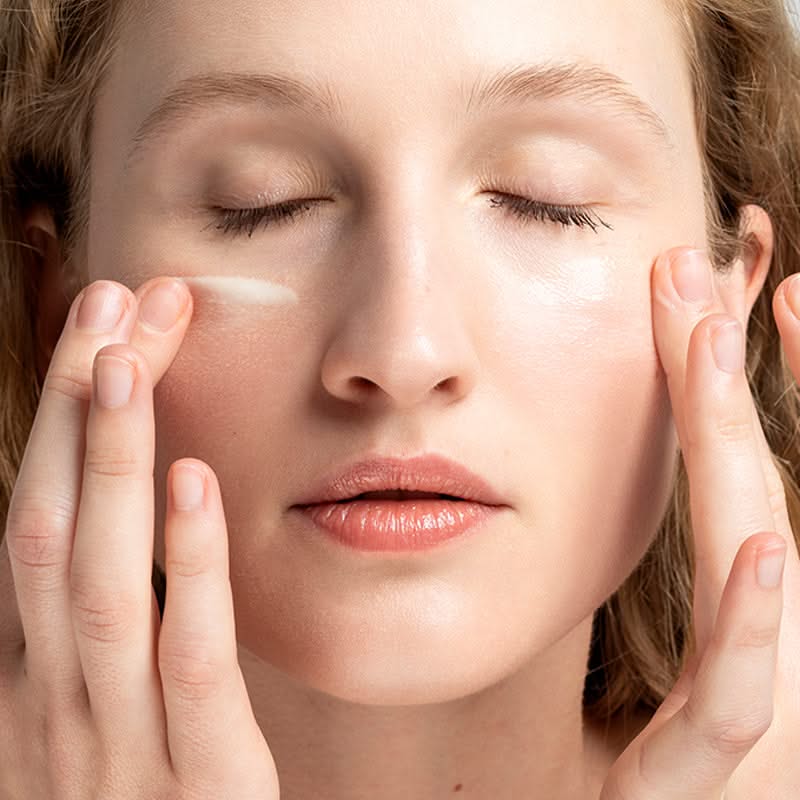 Frau mit geschlossenen Augen trägt Creme auf ihre Wangen auf.