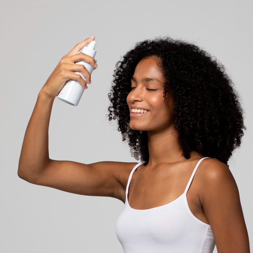 Frau mit lockigem Haar sprüht sich mit einem weißen Sprühflakon ins Gesicht und lächelt.