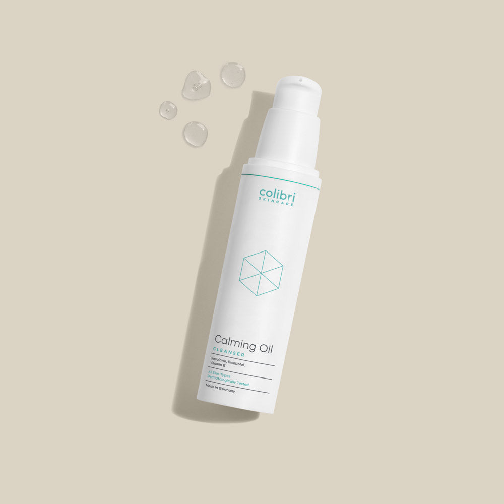 Flasche mit „Calming Oil Cleanser“ von Colibri Skincare, auf beigem Hintergrund mit Öltröpfchen daneben.