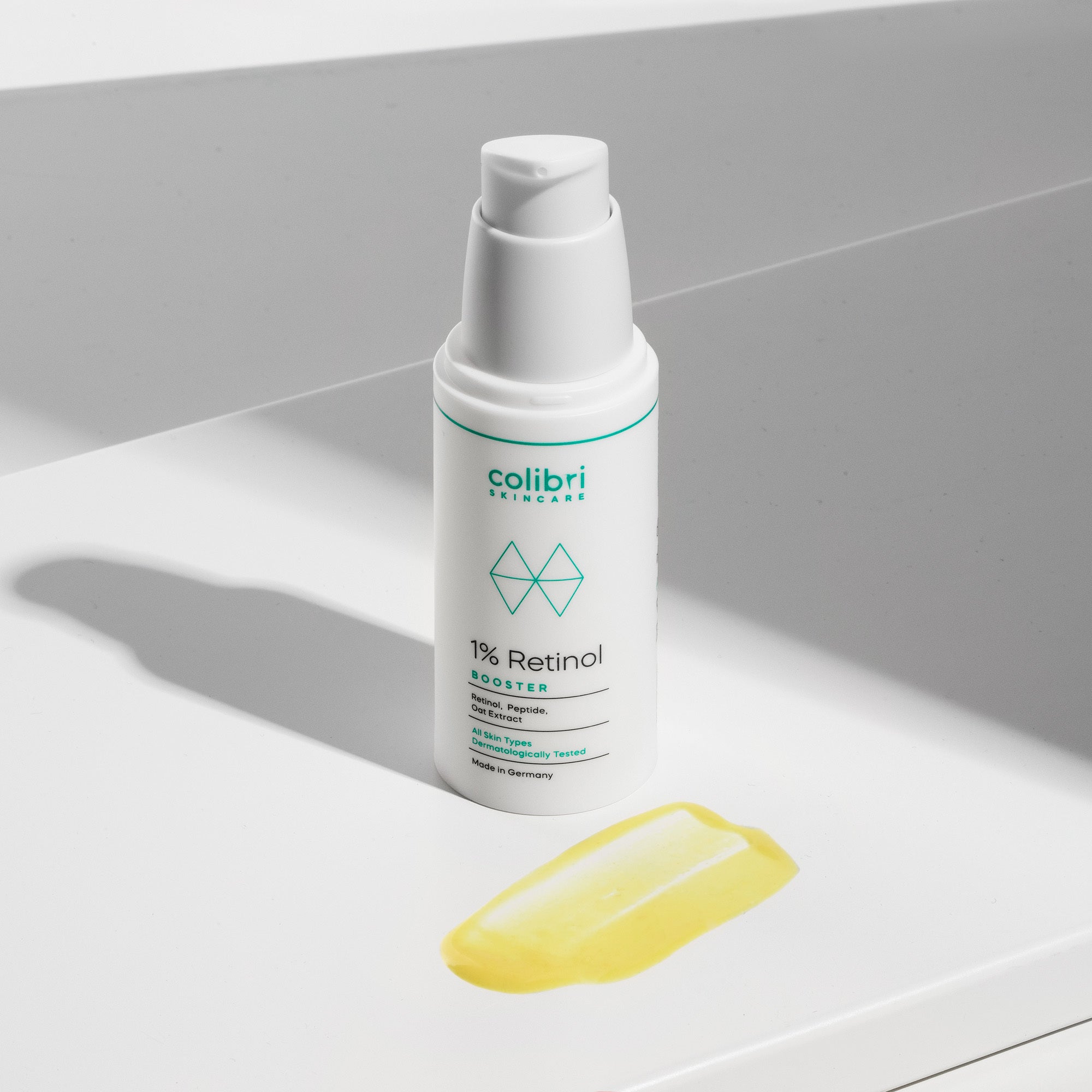 Flasche eines 1% Retinol-Boosters auf weißer Oberfläche neben einem gelben Tropfen des Produkts.