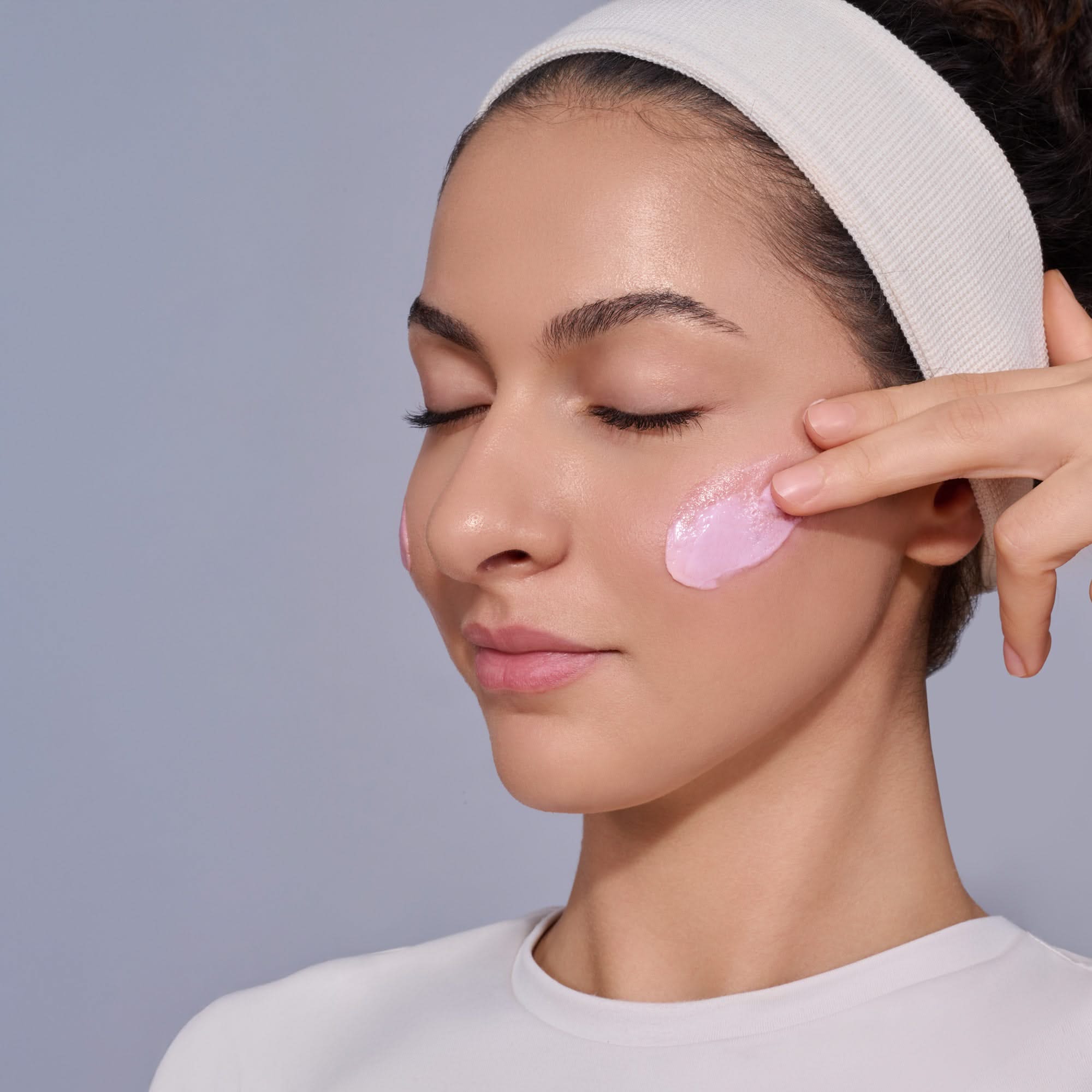 Frau trägt rosa Gesichtscreme auf ihre Wange auf und trägt ein Haarband.