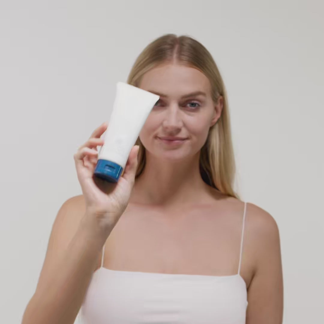 Eine Frau mit blonden Haaren hält eine weiße Tube mit blauer Kappe vor einem neutralen Hintergrund.