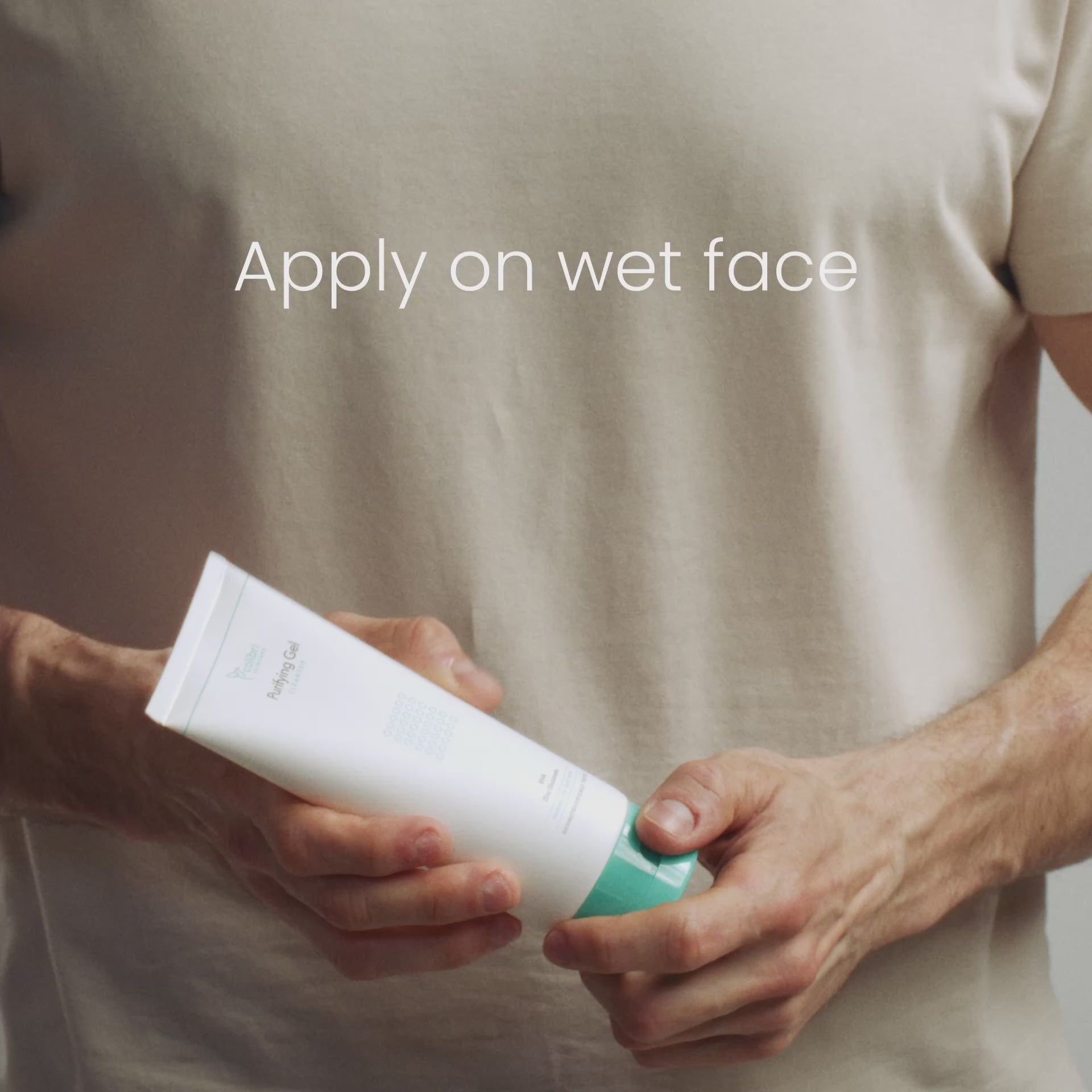 Ein Mann in einem beigen T-Shirt hält eine Tube Gesichtsreinigungsgel mit der Aufschrift „Apply on wet face“.