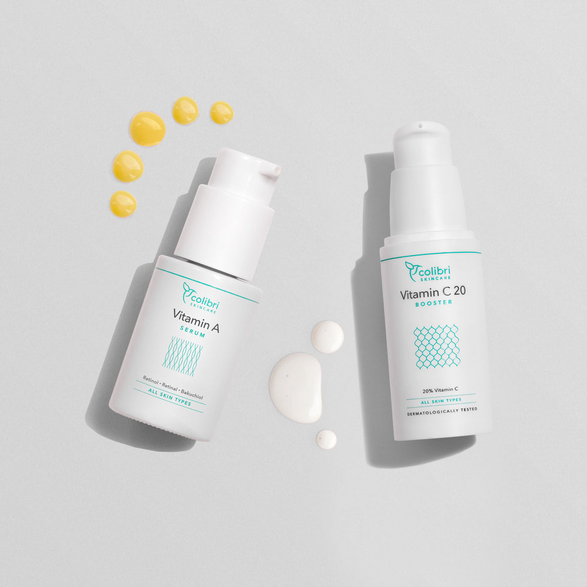 Zwei Fläschchen Hautserum mit Vitamin A und Vitamin C20, umgeben von kleinen Tropfen auf einem grauen Hintergrund.
