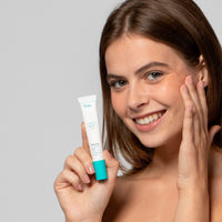 Frau lächelt und hält eine Tube Hautpflege, während sie das Produkt auf ihr Gesicht aufträgt.