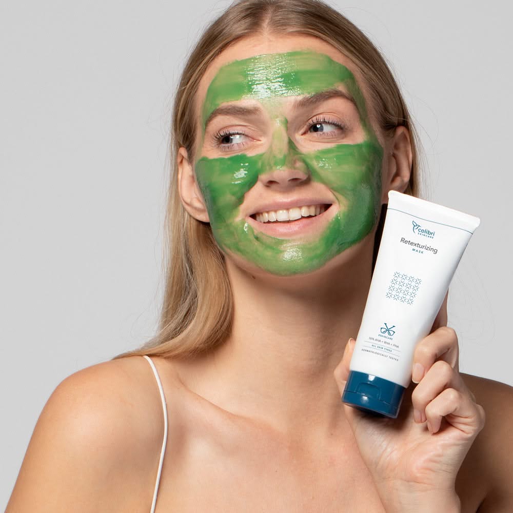 Frau mit grüner Gesichtsmaske hält eine Tube mit Hautpflegeprodukt in der Hand und lächelt.